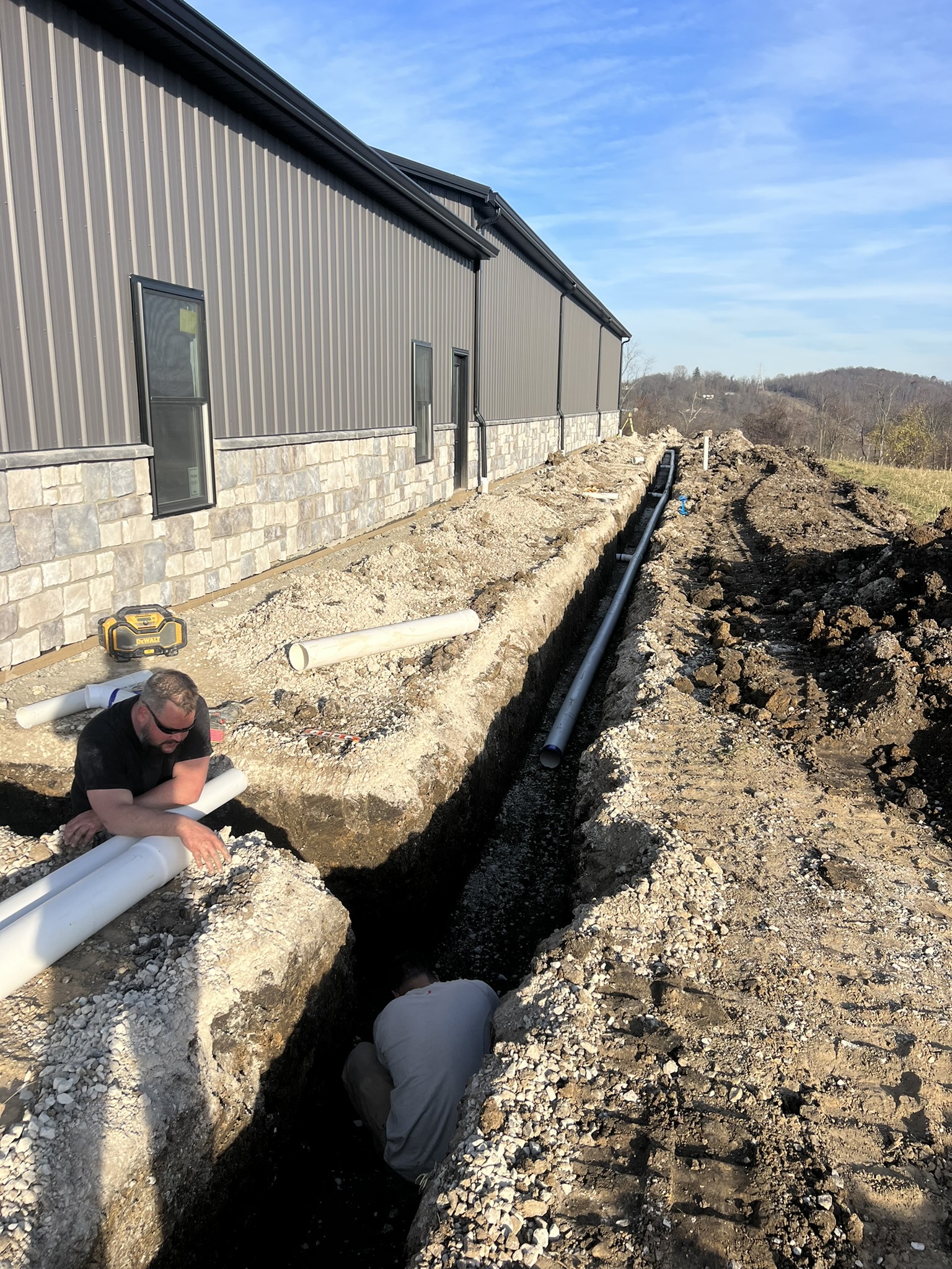 Two-man crew installing pipe in trench alongside barn